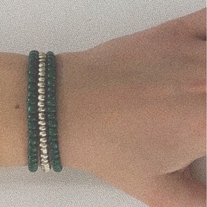 May include: A stack of three bracelets. The top and bottom bracelets are made of small, dark green beads. The middle bracelet is made of small, silver-colored beads. The bracelets are on a person's wrist.