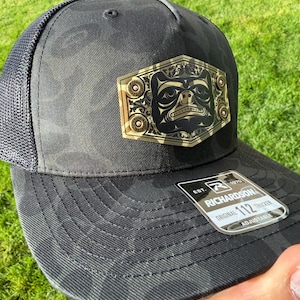May include: Black camouflage trucker hat with a mesh back. The front features a gold and black emblem with a tribal design. The hat has a curved brim and an adjustable strap. The tag reads "Richardson 112 Trucker".