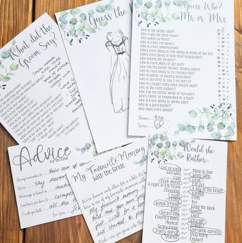 May include: A collection of bridal shower games on white cards with eucalyptus leaf accents. Games include "What Did the Groom Say?", "Guess the Bride", and "Would She Rather..." on a wooden surface.