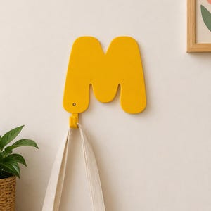 Op de afbeelding: Een felgele wandhaak in de vorm van de hoofdletter M. De haak heeft een glad, afgerond ontwerp met een kleine metalen schroef zichtbaar aan de linkerkant. Een bijpassende gele haak steekt aan de onderkant uit om voorwerpen aan op te hangen.