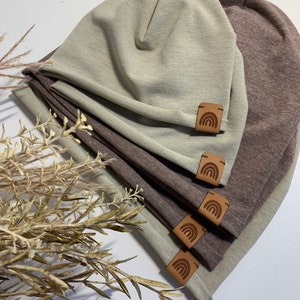 Könnte beinhalten: Ein Stapel von vier Mützen in Taupe- und Brauntönen. Jede Mütze hat ein kleines, braunes Lederetikett mit einem Regenbogen-Design. Die Mützen sind gefaltet und übereinander gestapelt. Getrocknete Pflanzen sind im Vordergrund.