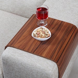 K&ouml;nnte beinhalten: Ein Holztablett mit abgerundeter Kante liegt auf einem hellgrauen Sofa. Das Tablett hat eine dunkelbraune Holzmaserung und enth&auml;lt eine kleine wei&szlig;e Schale mit gemischten N&uuml;ssen und ein Glas Rotwein.