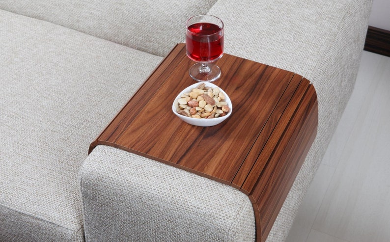 K&ouml;nnte beinhalten: Ein Holztablett mit abgerundeter Kante liegt auf einem hellgrauen Sofa. Das Tablett hat eine dunkelbraune Holzmaserung und enth&auml;lt eine kleine wei&szlig;e Schale mit gemischten N&uuml;ssen und ein Glas Rotwein.