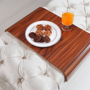 K&ouml;nnte beinhalten: Ein Holztablett mit abgerundeter Kante auf einem wei&szlig;en, gesteppten Ottoman. Auf dem Tablett befinden sich ein Teller mit Keksen und ein Glas Orangensaft.