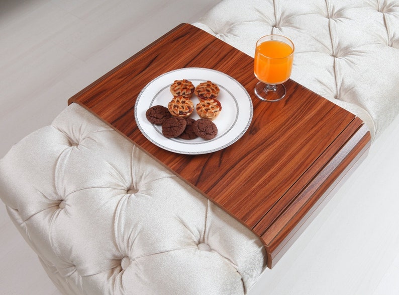 K&ouml;nnte beinhalten: Ein Holztablett mit abgerundeter Kante auf einem wei&szlig;en, gesteppten Ottoman. Auf dem Tablett befinden sich ein Teller mit Keksen und ein Glas Orangensaft.