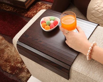 Könnte beinhalten: Ein dunkelbraunes Sofa-Armtablett aus Holz mit einer weißen Schale mit bunten Süßigkeiten und einem Glas Orangensaft. Das Tablett ist so konzipiert, dass es über eine Sofaarmlehne passt. Eine Person hält das Glas Saft.
