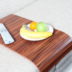 K&ouml;nnte beinhalten: Ein Tablett aus Holz mit brauner Oberfl&auml;che, mit einer Fernbedienung und einem Teller mit Obst. Das Tablett ist so konzipiert, dass es &uuml;ber die Armlehne eines Sofas oder Sessels passt.