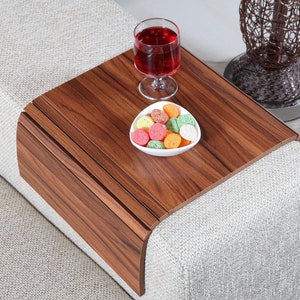 K&ouml;nnte beinhalten: Ein Holztablett mit abgerundeter Kante liegt auf einem wei&szlig;en Sofa. Das Tablett hat eine dunkle Holzmaserung und ist perfekt f&uuml;r Snacks und Getr&auml;nke. Ein Glas Rotwein und eine kleine wei&szlig;e Schale gef&uuml;llt mit bunten S&uuml;&szlig;igkeiten stehen auf dem Tablett.