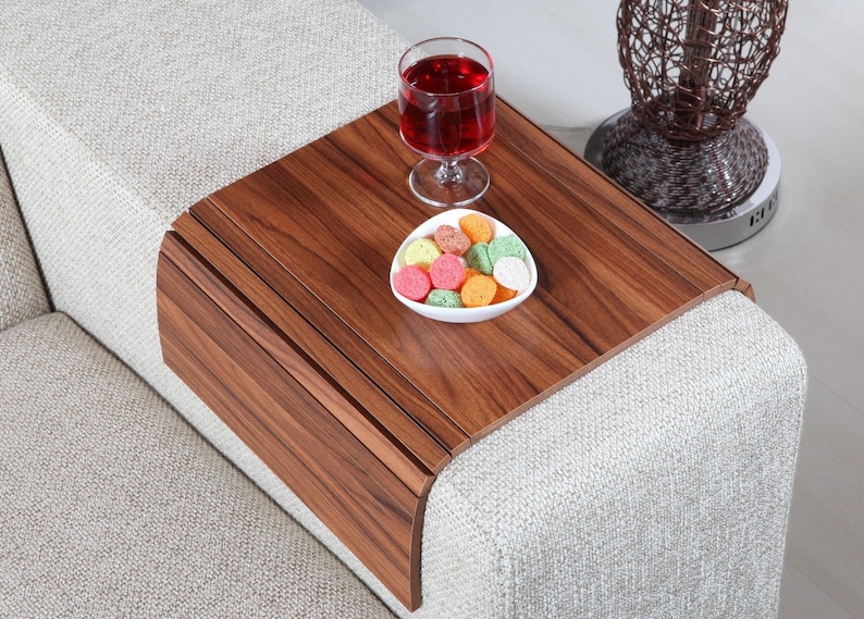 K&ouml;nnte beinhalten: Ein Holztablett mit abgerundeter Kante liegt auf einem wei&szlig;en Sofa. Das Tablett hat eine dunkle Holzmaserung und ist perfekt f&uuml;r Snacks und Getr&auml;nke. Ein Glas Rotwein und eine kleine wei&szlig;e Schale gef&uuml;llt mit bunten S&uuml;&szlig;igkeiten stehen auf dem Tablett.