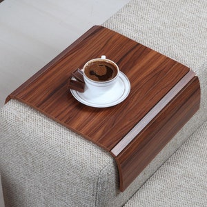 K&ouml;nnte beinhalten: Ein Holztablett mit abgerundeter Kante liegt auf der Armlehne eines Sofas. Auf dem Tablett stehen eine wei&szlig;e Tasse Kaffee und ein St&uuml;ck Schokolade.