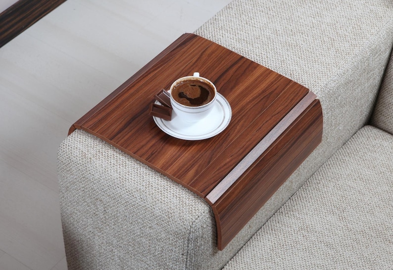 K&ouml;nnte beinhalten: Ein Holztablett mit abgerundeter Kante liegt auf der Armlehne eines Sofas. Auf dem Tablett stehen eine wei&szlig;e Tasse Kaffee und ein St&uuml;ck Schokolade.