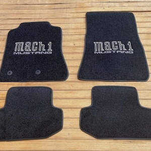 May include: Four black car floor mats. The front mats feature the text "Mach 1 Mustang" in white. The mats are made of a dark, textured material and are designed to fit a vehicle's floorboards.