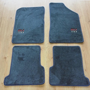 以下が含まれることがあります： 青い縁取りのある、濃い灰色の車のフロアマット4枚セット。フロントマットは独特の形状をしています。各マットには、赤と白で「GLI 16V」の刺繍が施されています。