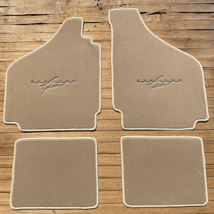 May include: Set of four beige car floor mats with white edging. The front mats have the word "Cadillac" embroidered in the centre.