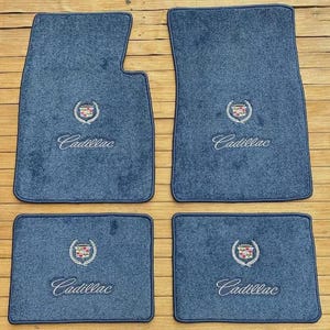 May include: Set of four blue car floor mats. Each mat features the Cadillac emblem embroidered in the center, with the Cadillac name below. The mats are made of a soft, textured material.