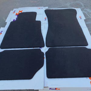 Puede incluir: Juego de cuatro alfombrillas negras para coche. Se muestran dos alfombrillas delanteras y dos traseras. Las alfombrillas son de un material oscuro, posiblemente carbón, sobre una superficie blanca.