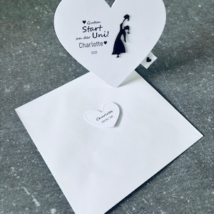Può includere: Un biglietto bianco a forma di cuore con la scritta "Guten Start an der Uni! Charlotte 2025" e la silhouette di una persona con la toga. Sono presenti anche una busta bianca e un piccolo adesivo a forma di cuore.