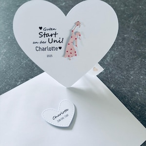 Puede incluir: Una tarjeta blanca en forma de corazón con el texto "Guten Start an der Uni! Charlotte 2025" y una pequeña ilustración de una persona con un vestido. También es visible una pegatina más pequeña en forma de corazón con el nombre "Charlotte".