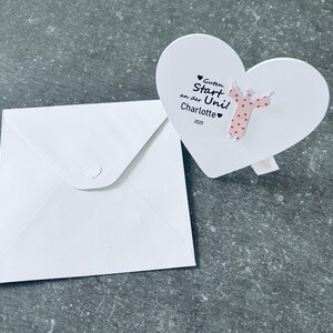 Puede incluir: Una tarjeta blanca en forma de corazón con el texto "Guten Start an der Uni! Charlotte 2025" y una figura de ángel rosa. También hay un sobre blanco con cierre en forma de corazón.