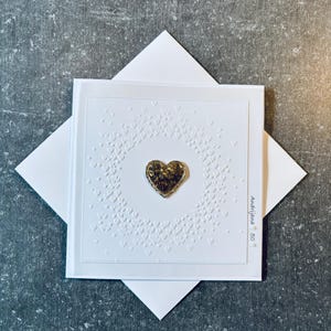Puede incluir: Una tarjeta de felicitación blanca con un corazón dorado en el centro, rodeado de un patrón de corazones en relieve. La tarjeta está colocada sobre un sobre blanco. El texto "Andrijana 50" está presente. Diseño sencillo.