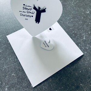 Puede incluir: Una tarjeta de felicitación blanca en forma de corazón con el texto alemán "Guten Start an der Uni! Charlotte 2025" y una silueta de una persona graduada. También se ve una pequeña etiqueta en forma de corazón con el nombre "Charlotte".