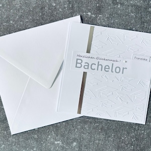 Op de afbeelding: Een witte afstudeerkaart en envelopset. De kaart heeft in reliëf gemaakte afstudeerhoeden en het woord "Bachelor" in grijs. De tekst "Herzlichen Glückwunsch! Franziska" is ook zichtbaar. Een verticale zilveren streep accentueert de kaart.