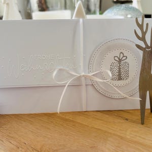 Op de afbeelding: Een witte kerstkaart met de in reliëf aangebrachte woorden "Frohe Weihnachten" en een cadeau-illustratie. De kaart is vastgebonden met een wit lint. Rechts staat een decoratief metalen rendier.