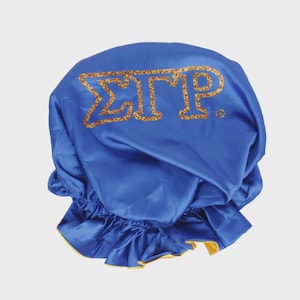 Könnte beinhalten: Eine königsblaue Satinhaube mit goldglitzernden Buchstaben, die "ΣΓΡ" buchstabieren. Die Haube hat einen gerüschten Rand mit goldenem Futter. Das Produkt dient zum Schutz der Haare.
