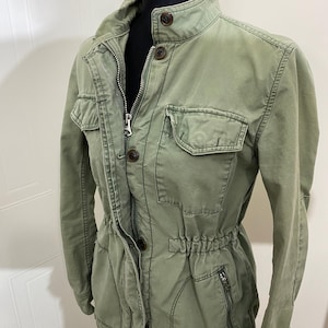 May include: An olive green utility jacket with a stand-up collar, multiple pockets, and a drawstring waist. The jacket features a zipper closure and button details. The fabric has a slightly worn appearance.