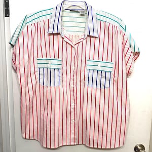 May include: A short-sleeved button-down shirt featuring a vertical striped pattern in red, white, blue, and green. The shirt includes a pointed collar, a front button placket, and two chest pockets with horizontal striped flaps in blue and green.