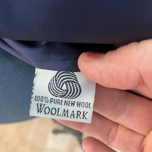 Peut inclure: Gros plan sur une &eacute;tiquette blanche avec le texte "100% PURE NEW WOOL WOOLMARK" et un symbole Woolmark. L'&eacute;tiquette est attach&eacute;e &agrave; un tissu bleu marine.