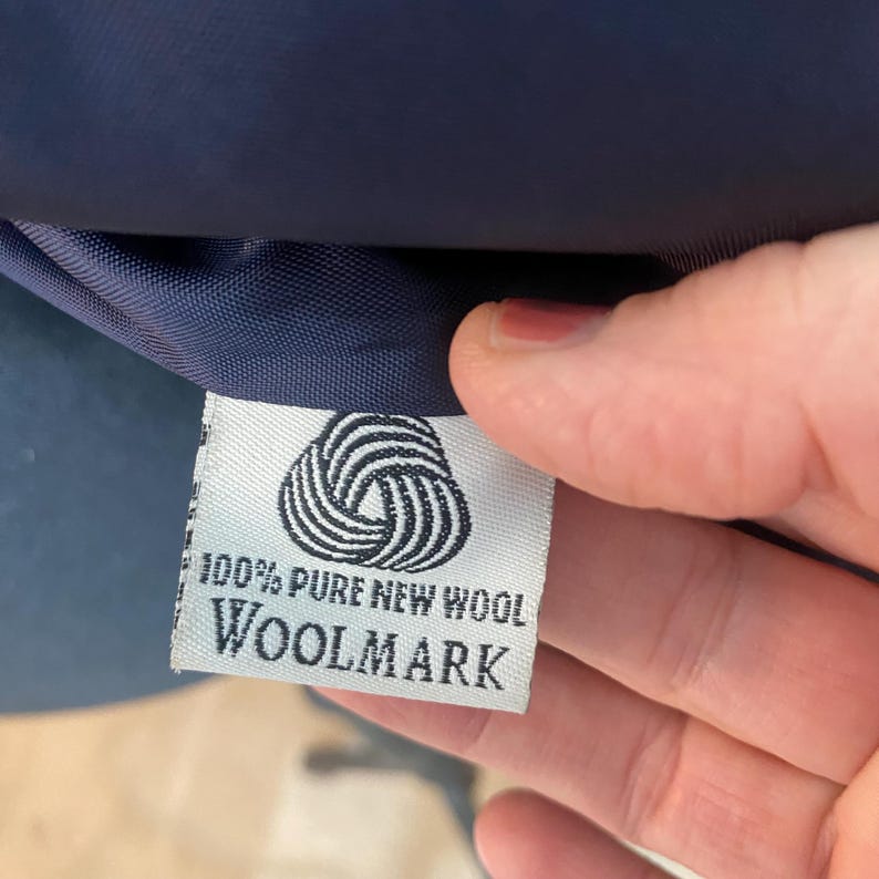 Peut inclure: Gros plan sur une &eacute;tiquette blanche avec le texte "100% PURE NEW WOOL WOOLMARK" et un symbole Woolmark. L'&eacute;tiquette est attach&eacute;e &agrave; un tissu bleu marine.