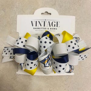 May include: A handmade hair bow featuring white, navy, and yellow ribbons. The bow has a polka dot pattern and a logo. The packaging reads "VINTAGE BARRETTES & BOWS INDIVIDUALLY HAND CRAFTED."