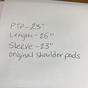 Peut inclure: Papier blanc avec des mesures manuscrites : PTP - 58 cm, Longueur - 64 cm, Manche - 58 cm, et "&eacute;paulettes originales". Le texte est &eacute;crit &agrave; l'encre noire.