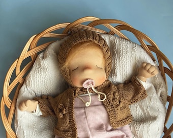 May include: A handmade cloth doll with blonde hair, wearing a brown knitted cardigan, pink overalls, and brown knitted booties. The doll is lying in a wicker basket with a white blanket.