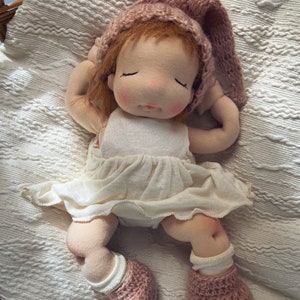 Pode incluir: Uma boneca de pano feita &agrave; m&atilde;o com um chap&eacute;u e botinhas de tric&ocirc; rosa, vestindo um vestido branco, deitada em um cobertor branco.