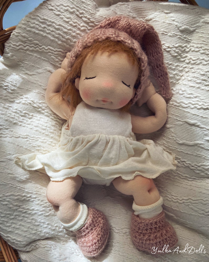 Pode incluir: Uma boneca de pano feita &agrave; m&atilde;o com um chap&eacute;u e botinhas de tric&ocirc; rosa, vestindo um vestido branco, deitada em um cobertor branco.