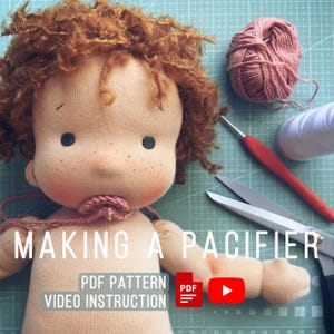 Puede incluir: Primer plano de una muñeca hecha a mano con cabello castaño rizado, que lleva un chupete rosa. La muñeca está tumbada sobre una estera de corte verde con tijeras, hilo y un ganchillo cerca. El texto "MAKING A PACIFIER" está escrito en letras blancas sobre fondo negro. El texto "PDF PATTERN VIDEO INSTRUCTION" también es visible.