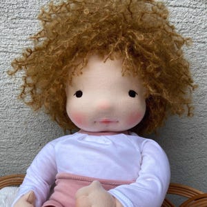 May include: A fabric doll with a round face, rosy cheeks, and a small mouth. The doll has voluminous, curly golden-brown hair and is wearing a white long-sleeved shirt paired with a dusty pink garment, featuring a soft, plush textile construction.