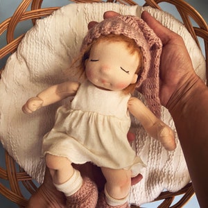 Pode incluir: Uma boneca de pano feita &agrave; m&atilde;o com um chap&eacute;u de malha rosa e botinhas combinando, vestindo um vestido branco, deitada em uma cesta de vime.
