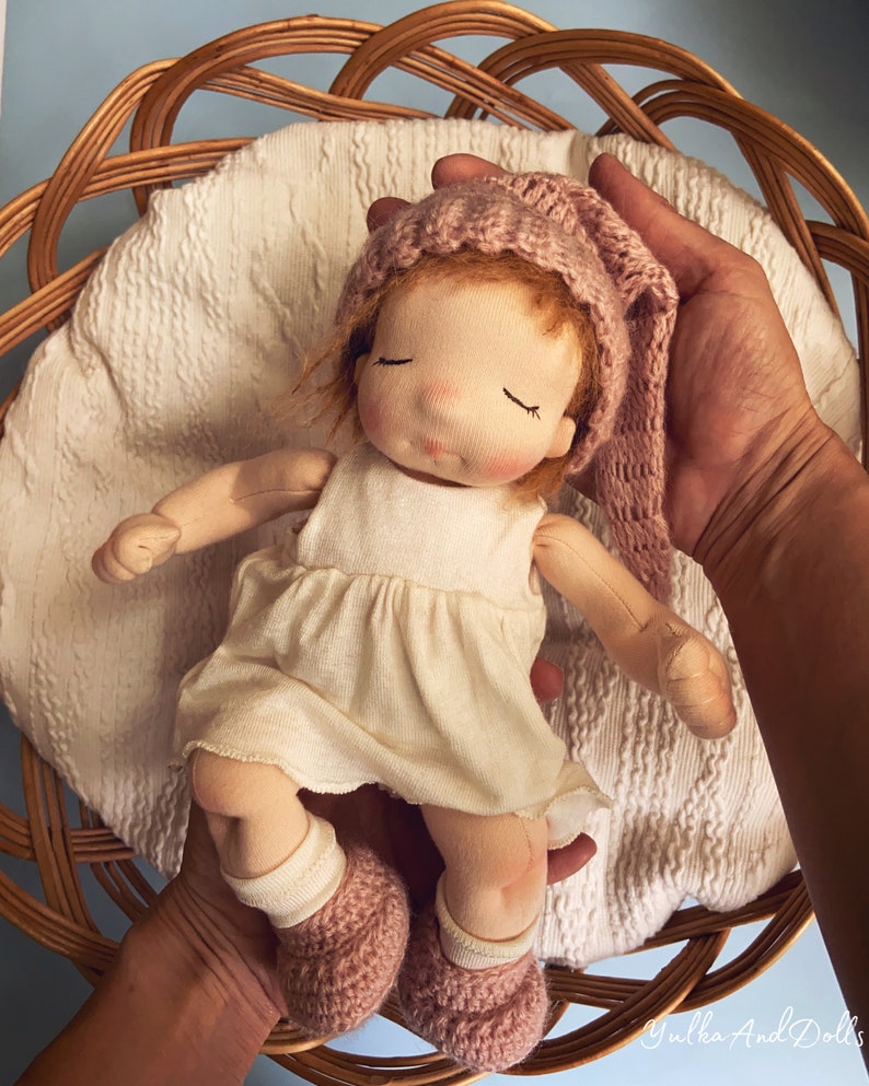 Pode incluir: Uma boneca de pano feita &agrave; m&atilde;o com um chap&eacute;u de malha rosa e botinhas combinando, vestindo um vestido branco, deitada em uma cesta de vime.