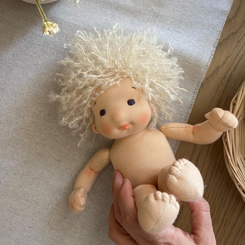 May include: A handmade cloth doll with a light peach skin tone, curly blonde hair, and rosy cheeks. The doll has simple facial features and is posed with arms and legs extended. The doll is approximately 8 inches tall.