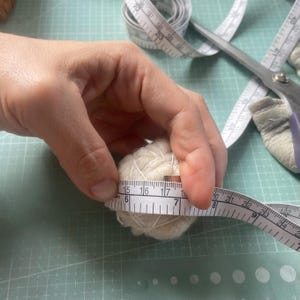 May include: A ball of cream-colored yarn is measured with a white measuring tape. The yarn ball is approximately 2 inches in diameter. Scissors and additional measuring tape are visible in the background, suggesting a crafting or sewing project.