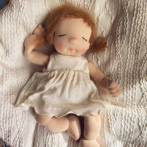 Pode incluir: Uma boneca de pano feita &agrave; m&atilde;o com cabelo castanho avermelhado, vestindo um vestido branco, est&aacute; deitada em um cobertor de croch&ecirc; branco. A boneca tem os olhos fechados e uma tez suave e rosada.