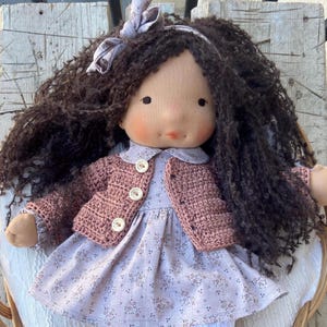 May include: A handmade doll with dark brown curly hair and a light complexion. The doll wears a light purple dress with a floral pattern and a crocheted pink jacket. A matching bow adorns the doll's hair. The doll is sitting on a white chair.