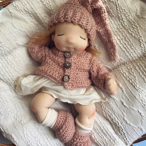 Pode incluir: Uma boneca de pano feita &agrave; m&atilde;o com um chap&eacute;u, um casaco e botinhas de malha rosa. A boneca est&aacute; deitada sobre um cobertor branco numa cesta de vime.