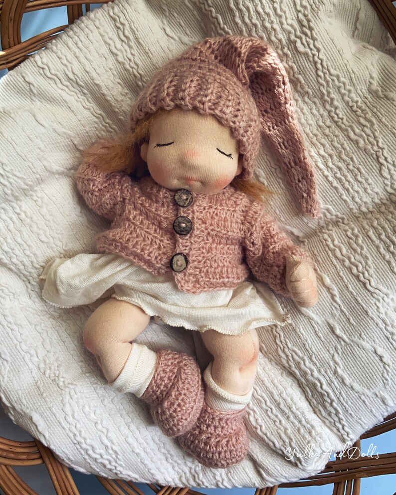 Pode incluir: Uma boneca de pano feita &agrave; m&atilde;o com um chap&eacute;u, um casaco e botinhas de malha rosa. A boneca est&aacute; deitada sobre um cobertor branco numa cesta de vime.