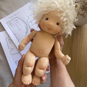 May include: A handmade cloth doll with a light skin tone and curly, light-colored hair. The doll has a simple, sweet face with rosy cheeks and small black eyes. The doll is being held, with sewing patterns visible in the background.