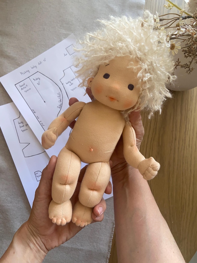 May include: A handmade cloth doll with a light skin tone and curly, light-colored hair. The doll has a simple, sweet face with rosy cheeks and small black eyes. The doll is being held, with sewing patterns visible in the background.