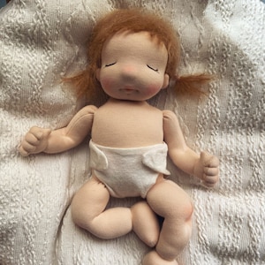 Pode incluir: Uma boneca de pano feita &agrave; m&atilde;o com uma fralda branca, cabelo castanho e olhos fechados, deitada em um cobertor de malha branca.